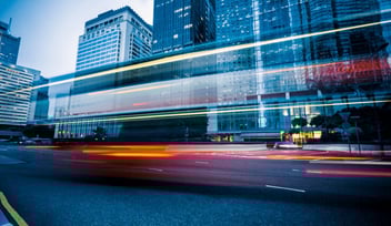 Image of bus lights wizzing passed in the foreground and skyscrapers in the background